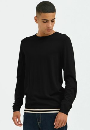 Chemise noire à manches longues avec un col rond, présentant une texture en maille côtelée et un ourlet rayé contrastant à la taille.