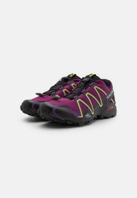 Tênis de trail running roxos e pretos com um cabedal texturizado, detalhes amarelos e um sistema de amarração r�ápida, apresentando uma sola robusta para tração.