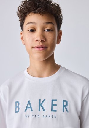 Young person with curly hair and freckles wearing a white T-shirt with blue "BAKER BY TED BAKER" text on a plain light background.
