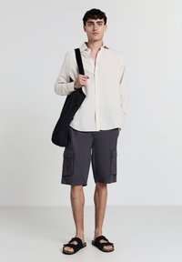 Chemise boutonnée beige clair, shorts cargo noirs avec deux poches latérales, sac fourre-tout noir et sandales noires avec une semelle texturée.