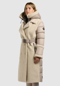 Beige mēteli ar teksturētu, pūkainu audumu priekšpusē un gludām, pūkām līdzīgām piedurknēm, plašu apkakli un jostu vidukļa daļā. Iekļautas kabatas ar rāvējslēdzējiem.