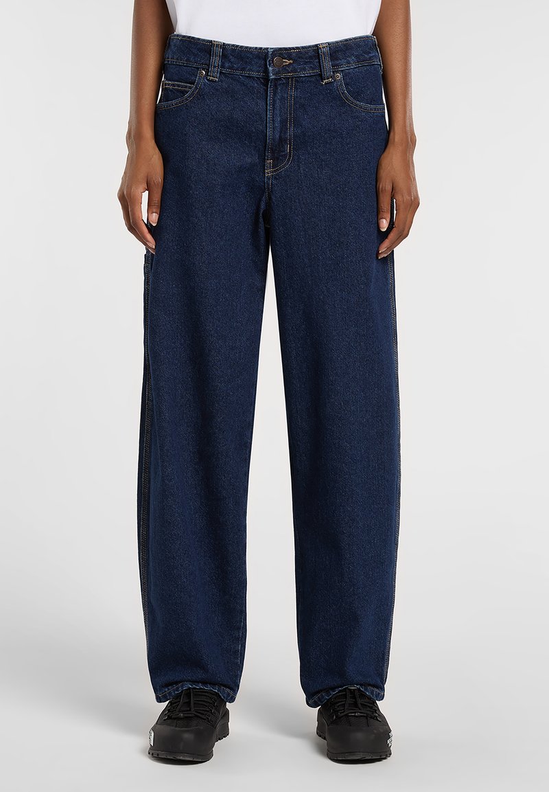 Mörkblå denimjeans med lös passform, enkelt knappstängning och detaljering vid sidoseam. Kombinerade med svart fotbeklädnad.