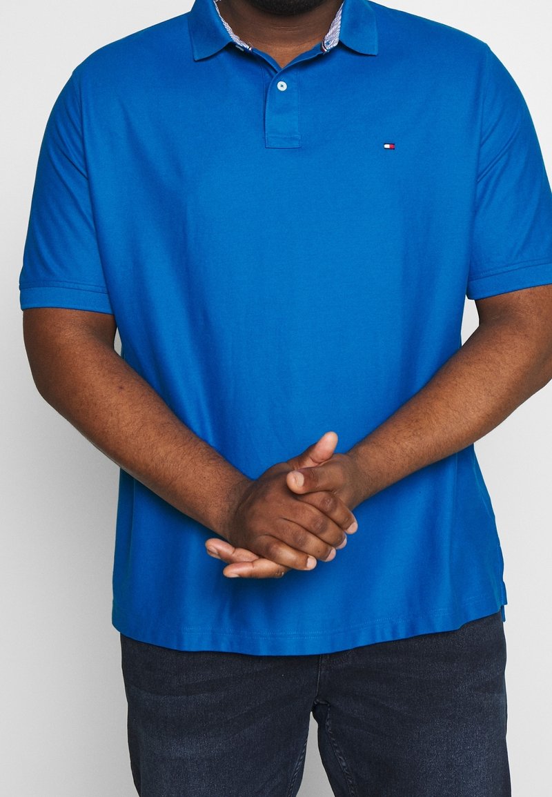 Blå polo skjorta med korta ärmar, med en krage och en diskret logotyp. Tyget verkar slätt med en enfärgad nyans och inga synliga mönster.