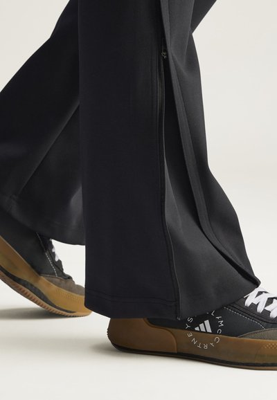 Schwarze Sporthose mit Seitenreißverschlüssen, kombiniert mit schwarz-grauen Turnschuhen mit Gummisohlen und weißen Schnürsenkeln, vor einem einfarbigen Hintergrund.