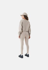 Sweat à manches longues beige coupé et leggings assortis en tissu doux, présentés de dos, associés à une casquette de couleur claire et des chaussures noires.