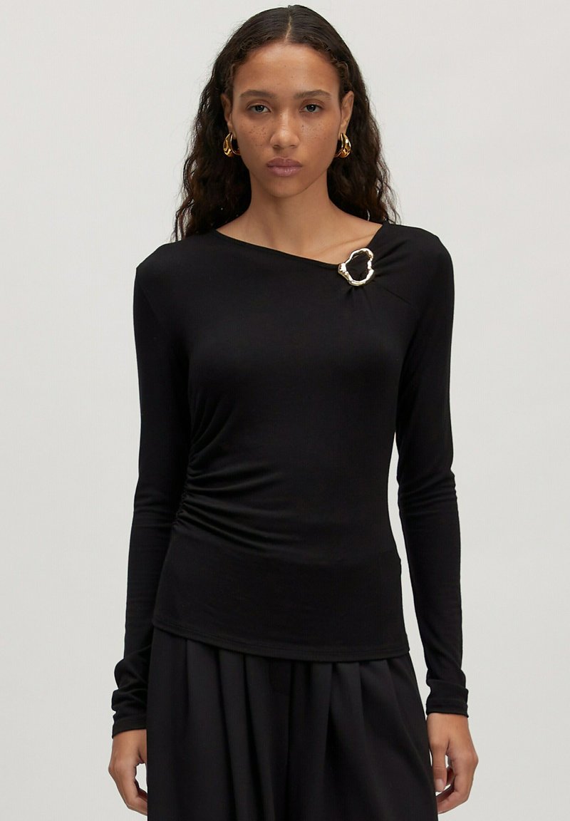 Schwarzes Langarmshirt mit drapiertem Design und einem silbernen Broschenakzent nahe der Schulter, aus glattem, dehnbarem Stoff.