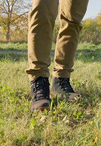 Czarne buty trekkingowe z teksturowanym materiałem, sznurowanym designem i gumowymi podeszwami, w połączeniu z beżowymi spodniami, stojące na trawiastym terenie.