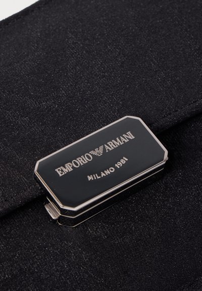 Черен правоъгълен метален етикет Emporio Armani със сребърни ръбове и текст, прикрепен към черна тъкан.