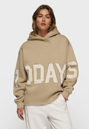 Felpa oversize beige con cappuccio e cordoncino, caratterizzata da grandi scritte bianche "100 DAYS". Il materiale sembra morbido con una finitura texturizzata.