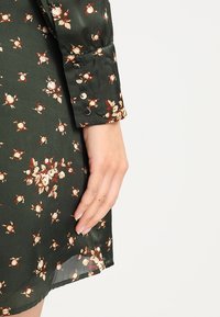 Robe florale verte avec une texture satinée ; présente de petites fleurs blanches et orange, un revers roulé avec des boutons et un ourlet transparent.