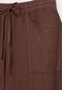 Pantalon marron à taille élastique avec cordon de serrage et grande poche avant à droite avec coutures visibles.