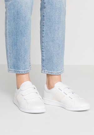 Białe sneakersy na rzepy noszone z jasnoniebieskimi dżinsami, pokazane od połowy łydki w dół na jednolitym tle.