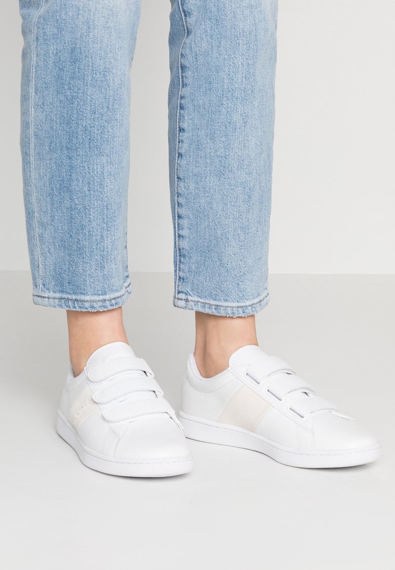 Baskets blanches à bride Velcro portées avec un jean bleu clair, montrées de la moitié du mollet vers le bas sur un fond uni.