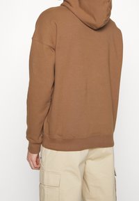 Sudadera con capucha marrón hecha de tela suave, con un ajuste relajado, puños acanalados y una etiqueta de logo visible en el lateral.
