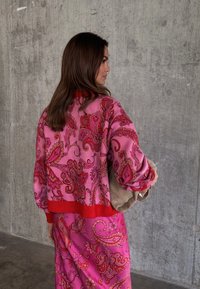 Γυναίκα με μακριά καστανά μαλλιά που φοράει ροζ και κόκκινη ενδυμασία με λουλουδάτο πασλέ σχέδιο, κρατώντας μια μπεζ τσάντα, στέκεται μπροστά σε τσιμεντένιο τοίχο.