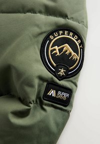 Olivengrønt stof med en fyldig tekstur, der har to broderede lapper: et cirkulært logo og et rektangulært mærke med guld accenter.