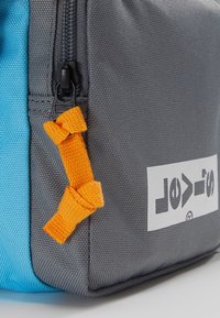 Grå og blå stofpose med en struktureret overflade, der har en orange lynlås-træk og et hvidt logo på siden.