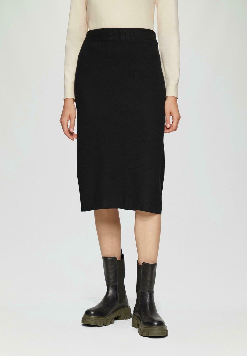 Femme portant une jupe noire longueur genou, un haut beige à manches longues et des bottes en cuir noires à semelle épaisse, debout les mains détendues.