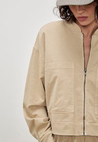 Vrouw in een beige zip-up jas met grote voorzakken en een geruite wollen pet, kijkend naar beneden.