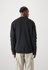 Felpa nera con collo rotondo e maniche lunghe, caratterizzata da una toppa tessuta blu navy sulla manica sinistra, realizzata in morbido tessuto di cotone.