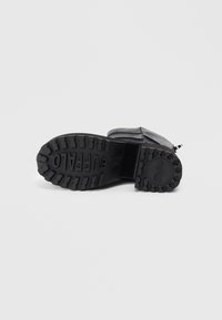 Bota negra tumbada de lado mostrando la suela texturizada con el logo "BUFFALO" y un tacón grueso sobre un fondo blanco liso.