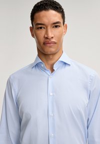 Camicia azzurra chiara con colletto strutturato, maniche lunghe e chiusura con bottoni. Il tessuto sembra liscio con una sottile texture e un leggero lucido.