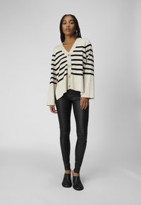 Beige og sort stribet cardigan med V-hals, med knapper ned langs midten og brede ærmer, parret med sorte imitation læder bukser.