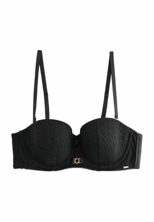 Femme portant un soutien-gorge noir sans bretelles et un sous-vêtement taille haute assorti à bordure en dentelle, posant en intérieur avec la main sur la hanche.