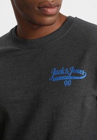 Tmavě šedá mikina s kulatým výstřihem a modrým logem "Jack & Jones Originals 90" na levé straně hrudníku.