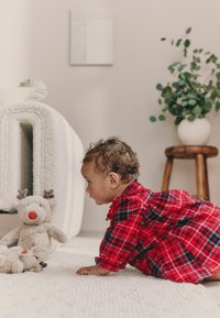 Rød ternet kjole, der minder om en tunika, blødt stof med fine flæser. Baby kravler på et neutralt struktureret tæppe og kigger mod et blødt rensdyr legetøj.