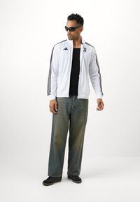 Hvid lynlåsjakke med sorte striber, sort skjorte, baggy lyseblå jeans og mørke sko; iført solbriller og ringe.