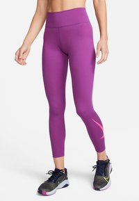 Fioletowe legginsy z wysokim stanem i gładką fakturą, z różowym logo Swoosh z boku, noszone z ciemnymi butami sportowymi.