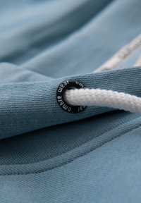 Jasnofioletowa tkanina o teksturowej powierzchni, z czarnym otworem i białym sznurkiem do ściągania. Widoczne detale szwów wzdłuż krawędzi.