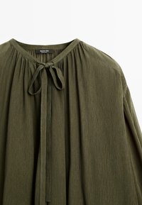 Blusa verde scuro realizzata in tessuto testurizzato, dotata di scollatura rotonda, dettagli plissettati e chiusura con cravatta al collo.