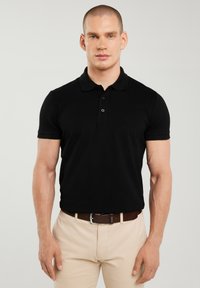 Mann trägt schwarzes Poloshirt mit kurzen Ärmeln und beige Hose mit braunem Gürtel, steht vor hellem einfarbigem Hintergrund.