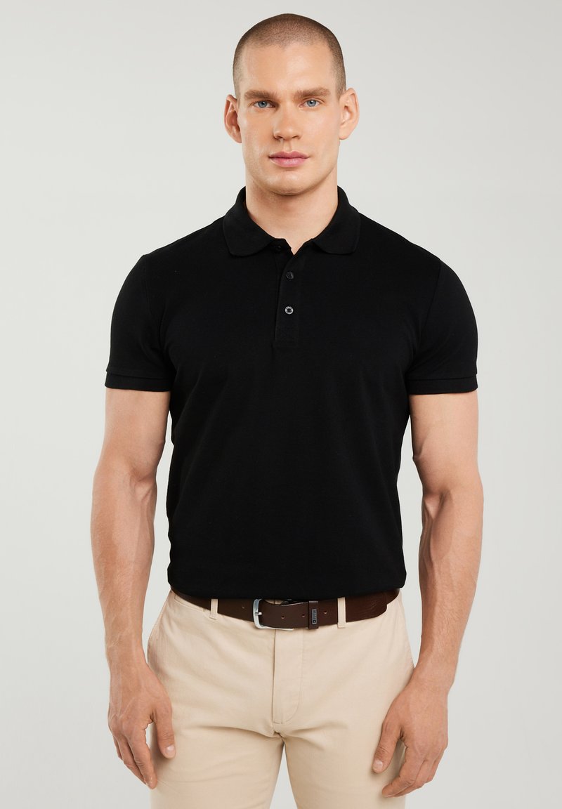 Mann trägt schwarzes Poloshirt mit kurzen Ärmeln und beige Hose mit braunem Gürtel, steht vor hellem einfarbigem Hintergrund.