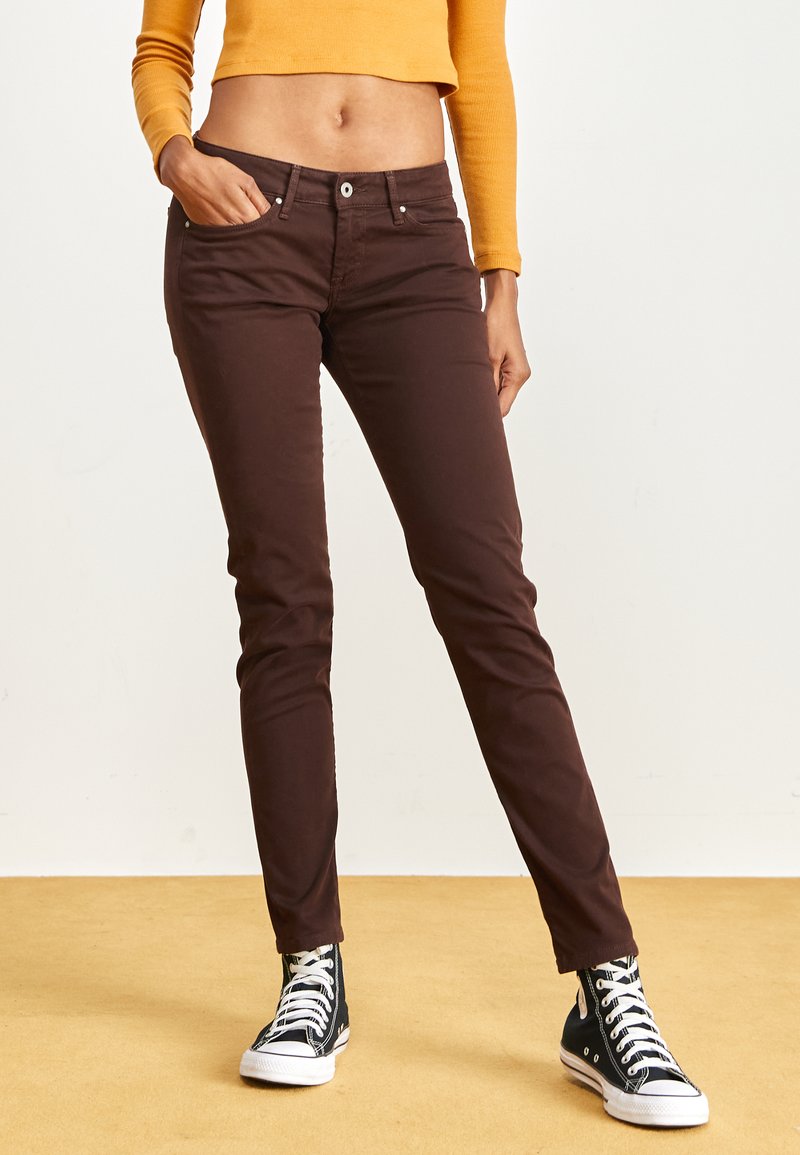 Braune Slim-Fit-Hosen aus Baumwolle mit glatter Textur, mit einer Fronttasche, Gürtelschlaufen und einem Knopfverschluss. Dazu werden schwarze High-Top-Sneaker getragen.