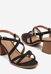 Sandales à talons noirs avec un design à lanières croisées, en tissu texturé, bout ouvert et un talon bloc moyen. Fermeture à boucle au niveau de la cheville.