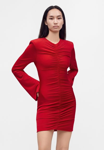 Femme aux longs cheveux foncés portant une robe rouge ajustée avec un centre froncé et de longues manches évasées, debout les mains sur les hanches sur fond blanc.