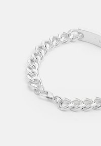 Pulseira de prata com elos de corrente entrelaçados, textura suave e fecho seguro. Inclui um detalhe metálico plano gravado com o nome da marca.