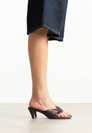 Pied gauche portant une mule à talon ouvert marron foncé avec une bride torsadée et un talon bloc court, associée à un pantalon court en jean foncé.