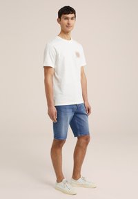 Bela bombažna majica s manjšo grafiko na prsih, v kombinaciji z modrimi denim kratkimi hlačami in belimi tenis škornji. Enostaven dizajn, sproščen kroj.