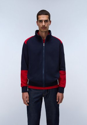 Mann mit einer marineblauen und roten Zip-Jacke und dunklen Jeans steht vor einem schlichten, hellen Hintergrund und blickt mit neutralem Gesichtsausdruck nach vorn.