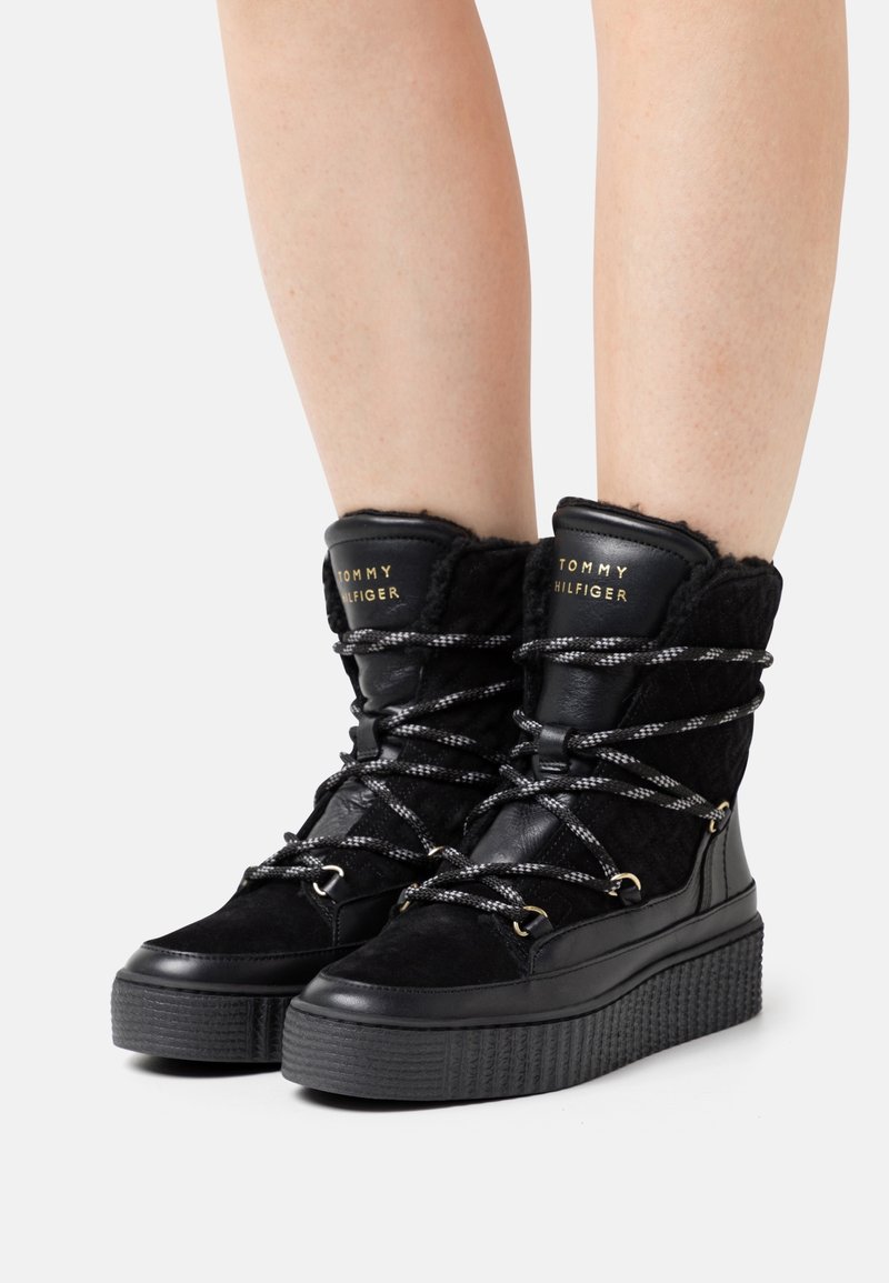Bottes d'hiver noires Tommy Hilfiger avec semelles épaisses et lacets à motifs, portées sur des jambes nues sur fond blanc.