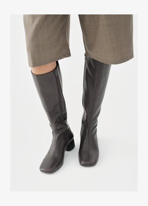 Braune kniehohe Lederstiefel mit schlankem Design, eckigen Zehen und Seitenreißverschlüssen, kombiniert mit hellgrauen strukturierten Hosen.