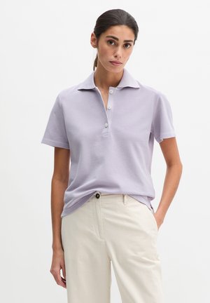 Kobieta o ciemnych włosach związanych w kucyk, ubrana w jasnofioletową koszulę z krótkim rękawem z kołnierzykiem oraz jasnobeżowe spodnie, stojąca z jedną ręką w kieszeni.