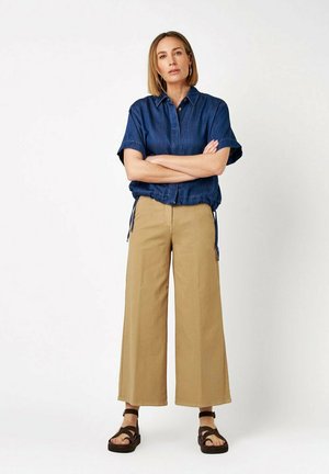 Donna in piedi con le braccia incrociate che indossa una camicia blu a maniche corte, pantaloni beige a gamba larga e sandali neri su sfondo bianco.