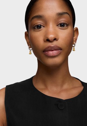 Top negro sin mangas con escote redondo y detalle de botón. La modelo lleva pendientes de aro plateados con un detalle dorado. Textura de piel suave.