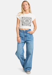 T-shirt bianca cropped con design grafico nero, abbinata a jeans a gamba larga di colore azzurro chiaro. Il modello è in piedi con le mani sui fianchi, indossando sneakers.