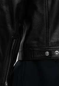 Μαύρο δερμάτινο μπουφάν με ορατό φερμουάρ στο μανίκι, λεπτομέρεια με κουμπωτό ιμάντα στη μέση και ραμμένη υφή στην λεία επιφάνεια του υφάσματος.